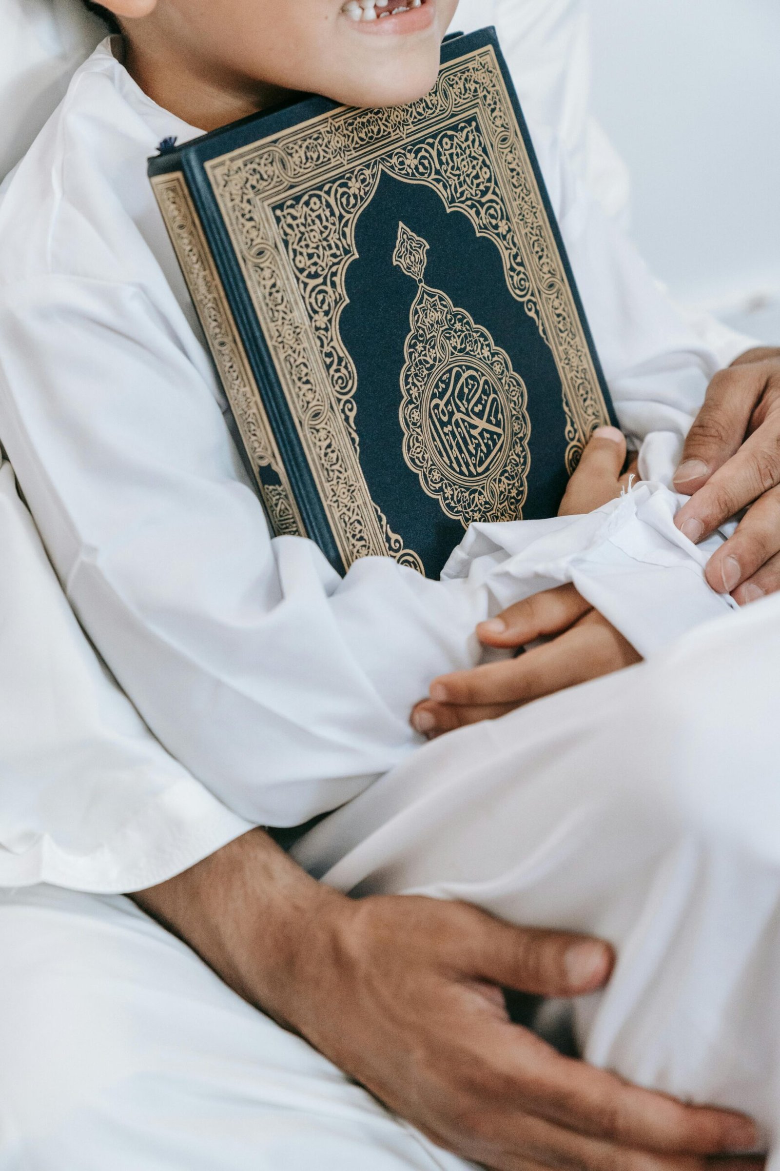 A child wearing a thobe embraces a Quran, capturing a warm and intimate moment indoors.